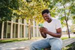 happy african american young guy using his cellphone at university college ,sending text messages sitting on a bench outdoors. Portrait of student man watching the screen of his mobile phone at campus