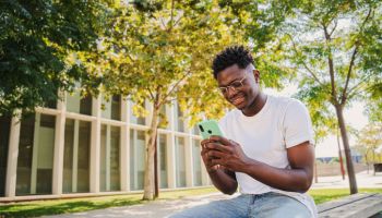happy african american young guy using his cellphone at university college ,sending text messages sitting on a bench outdoors. Portrait of student man watching the screen of his mobile phone at campus