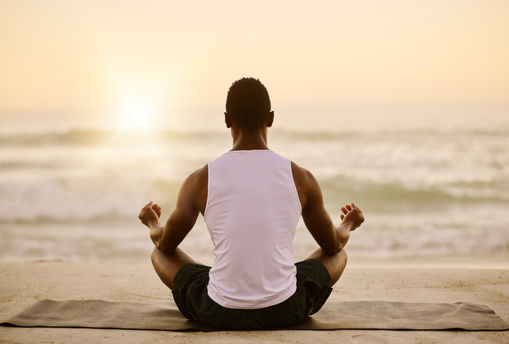 Relaxed, healthy and zen man meditating on beach in lotus pose by sea or ocean on quiet morning. Back of calm and spiritual yogi breathing, finding mental balance or wellness in holistic sunrise yoga