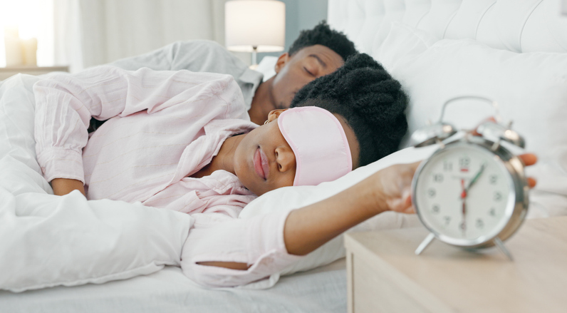 Woman, sleeping and snooze alarm in bed with fatigue, tired or lazy in morning with man in home. Black couple, girl stop clock and rest in bedroom, house or annoyed in mask at wake up time with dream