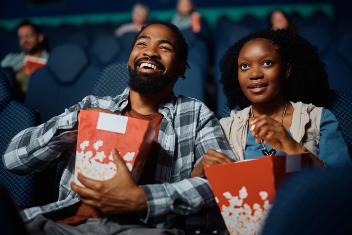 Happy black couple eating popcorn while watching movie in theater.