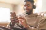 Happy man in brown hoodie using phone, studying, learning, shopping, working online, talking, listening to music, audiobook, podcast while sitting on a safa at home Happy time at home concept