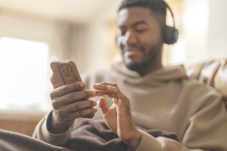 Happy man in brown hoodie using phone, studying, learning, shopping, working online, talking, listening to music, audiobook, podcast while sitting on a safa at home Happy time at home concept