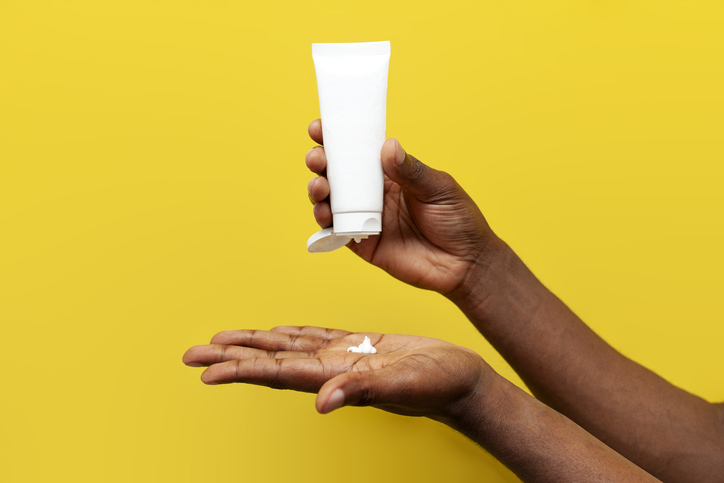 sunscreens for melanated skin - hands of african american man hold white empty tube of cream on yellow isolated background, hands squeeze out