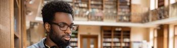 Dark-skinned young man in eyeglasses with a book in hands in the library