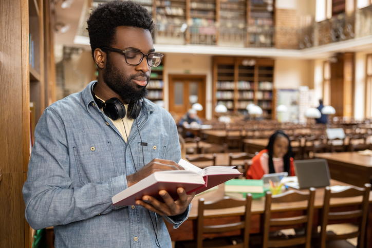 books by christian authors - Dark-skinned young man in eyeglasses with a book in hands in the library