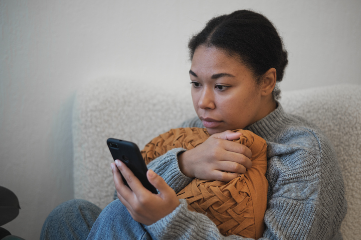 Social media depression. Sad upset African American woman holding phone reading sms message receiving bad news, disappointed black female sitting on bed looking at smartphone screen scrolling social media feeling depressed