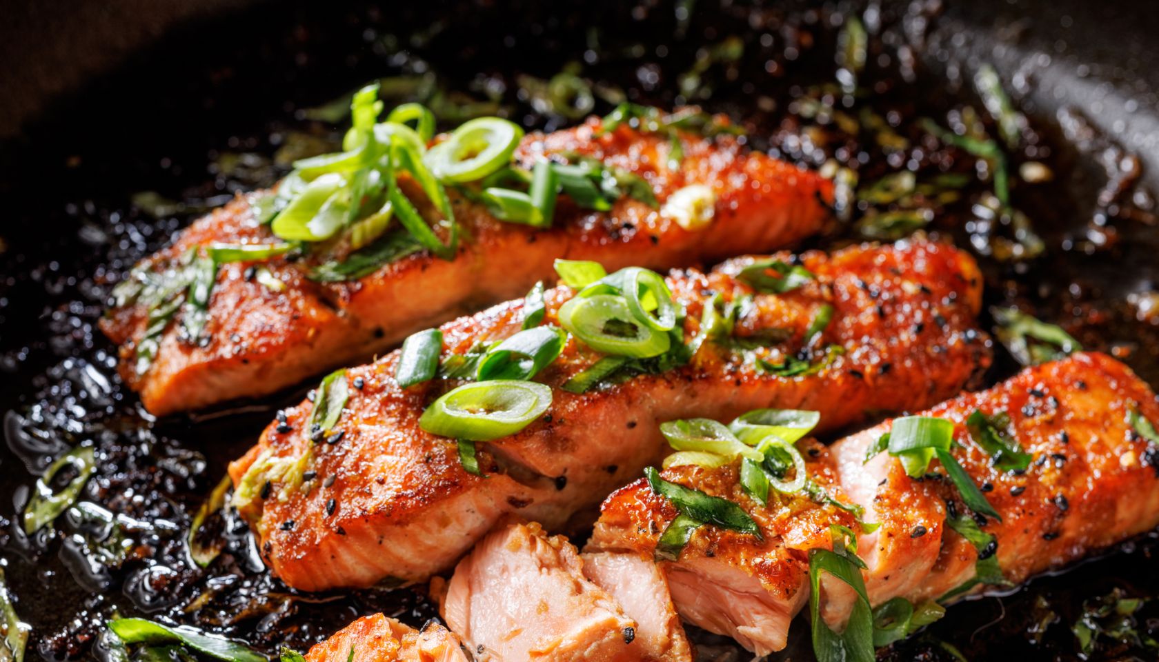 Close-up view of teriyaki salmon sprinkled with chopped scallion in a black baking dish