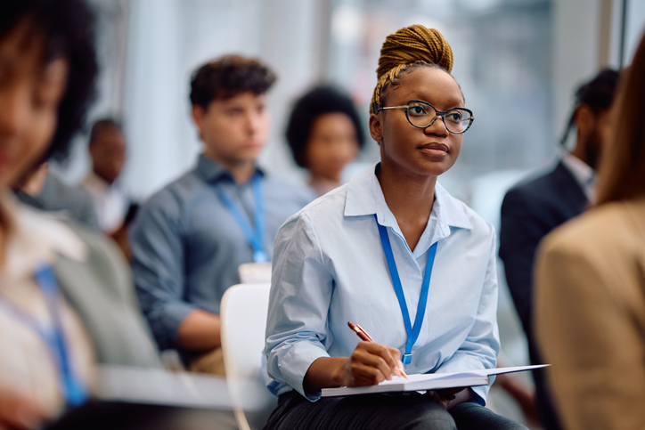 Black female manager writing in notebook while attending business seminar in conference hall.